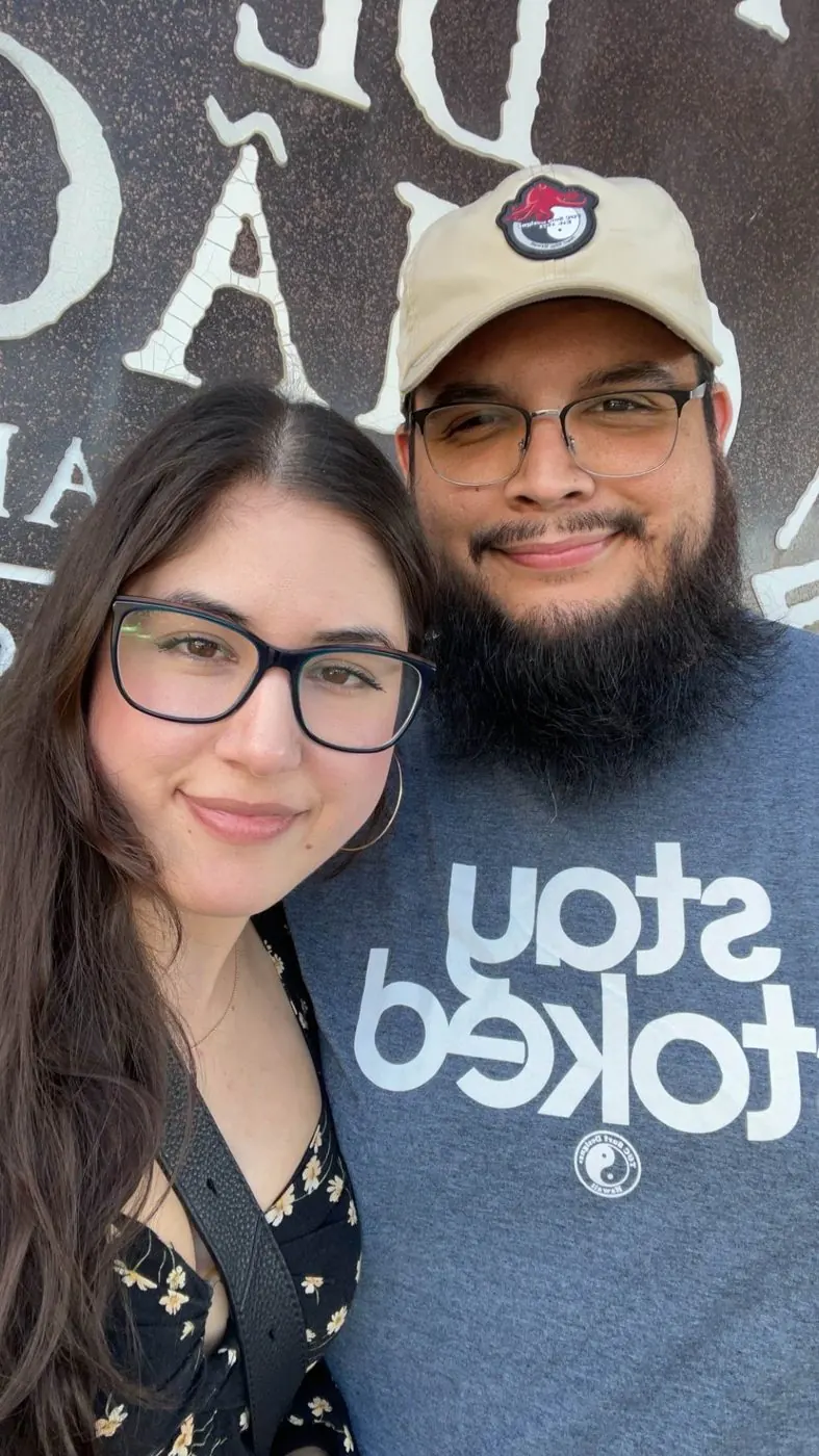 Brandon and his wife, close together, smiling, in front of a big painted sign.