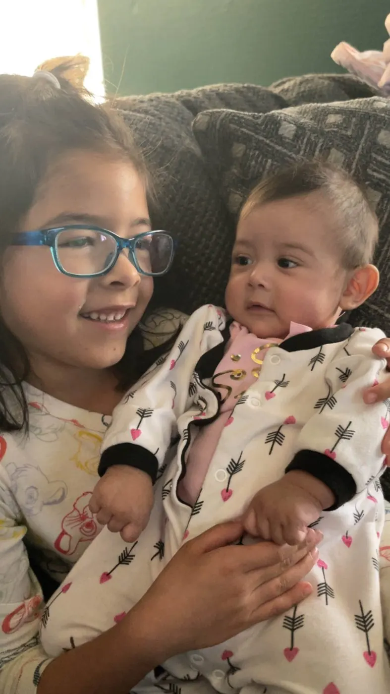 Brandon's older daughter on the couch, holding the baby, someone else's forearm under the baby and fingers curled round her side.