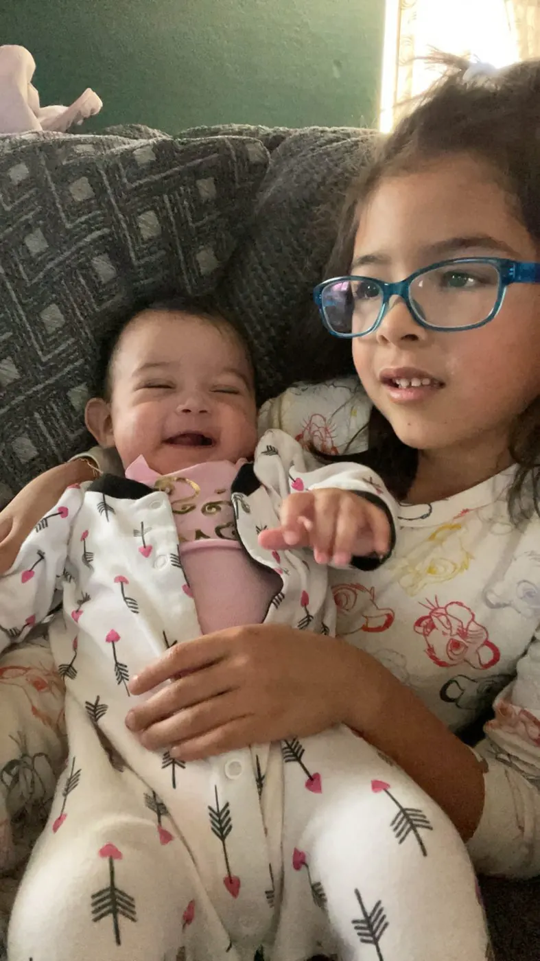 The baby laughing on the couch, her own arm raised and pointing at the camera, held by her older sister, someone else's forearm behind the baby at the left edge.