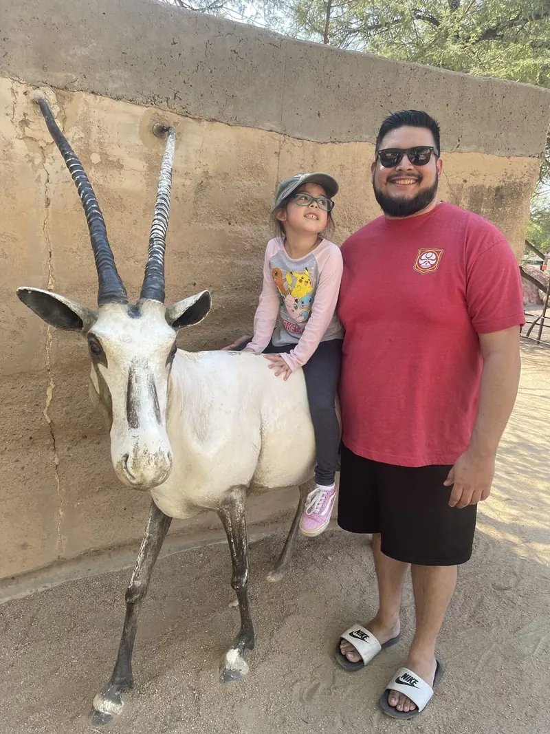 Brandon's daughter riding a statue of an oryx while he stands beside it, grinning in sunglasses.