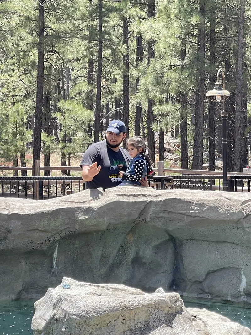 Brandon holding his daughter behind a large rock wall, a railing behind them and the pond below, one hand raised in a wave, looking at the camera.