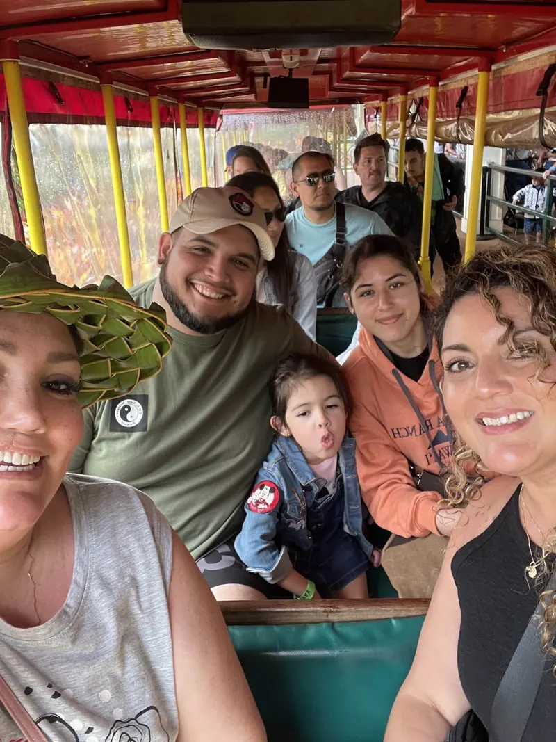 The whole family in a red open-sided tram with other passengers, the child between the adults pursing her lips and looking away.