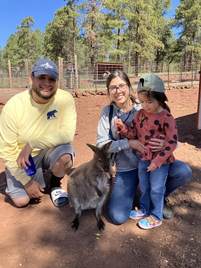 Brandon's wife kneeling in the pines, a wallaby taking something orange from her hand, Brandon on one knee beside her, their daughter standing against her.